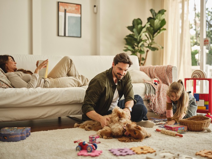 Family spending time together at home, with adults relaxing and a child playing on the floor beside a dog in a living room.