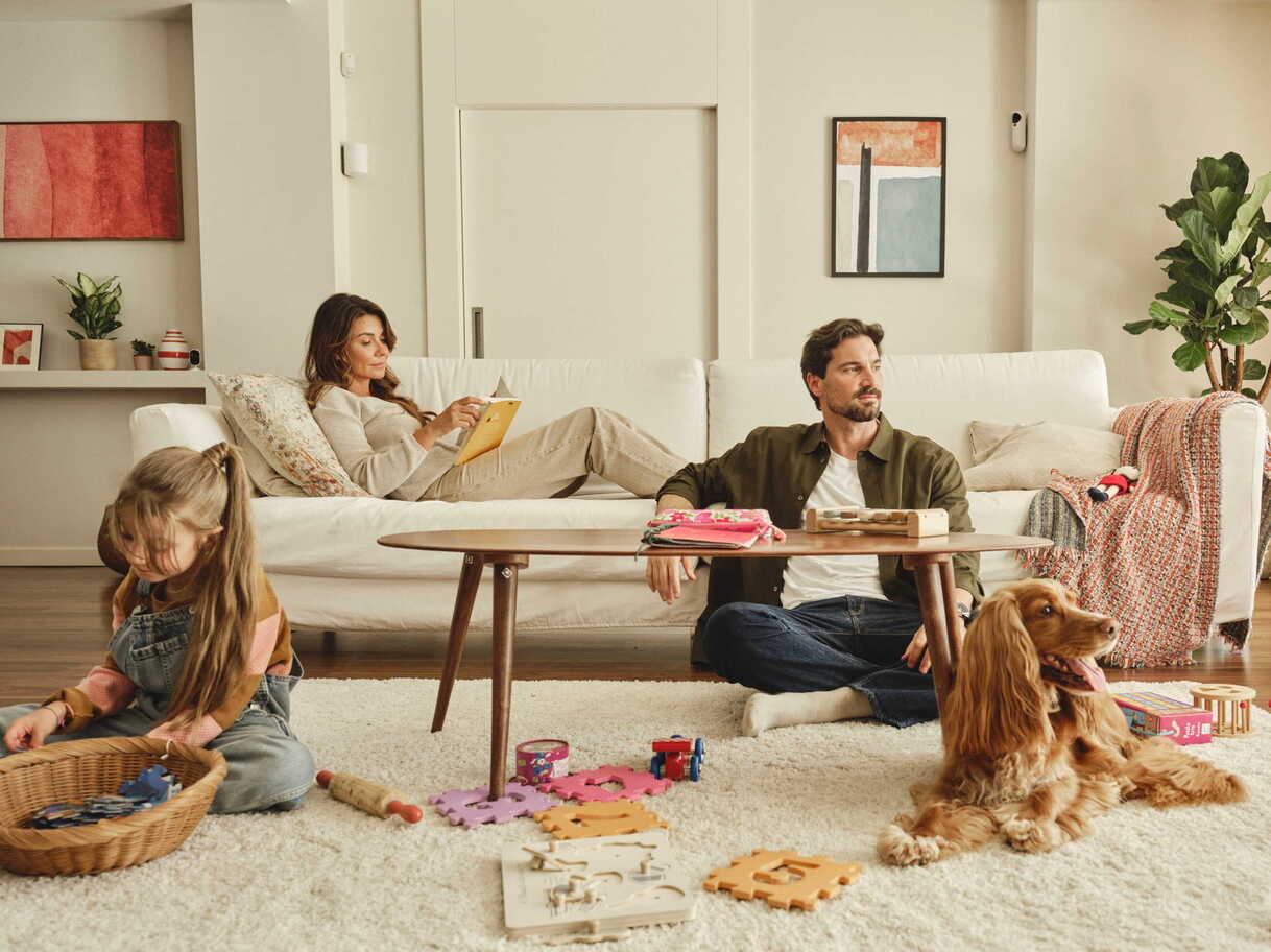 Family spending time together in a living room, with a child playing on the floor, a dog resting nearby, and two adults relaxing on a sofa.