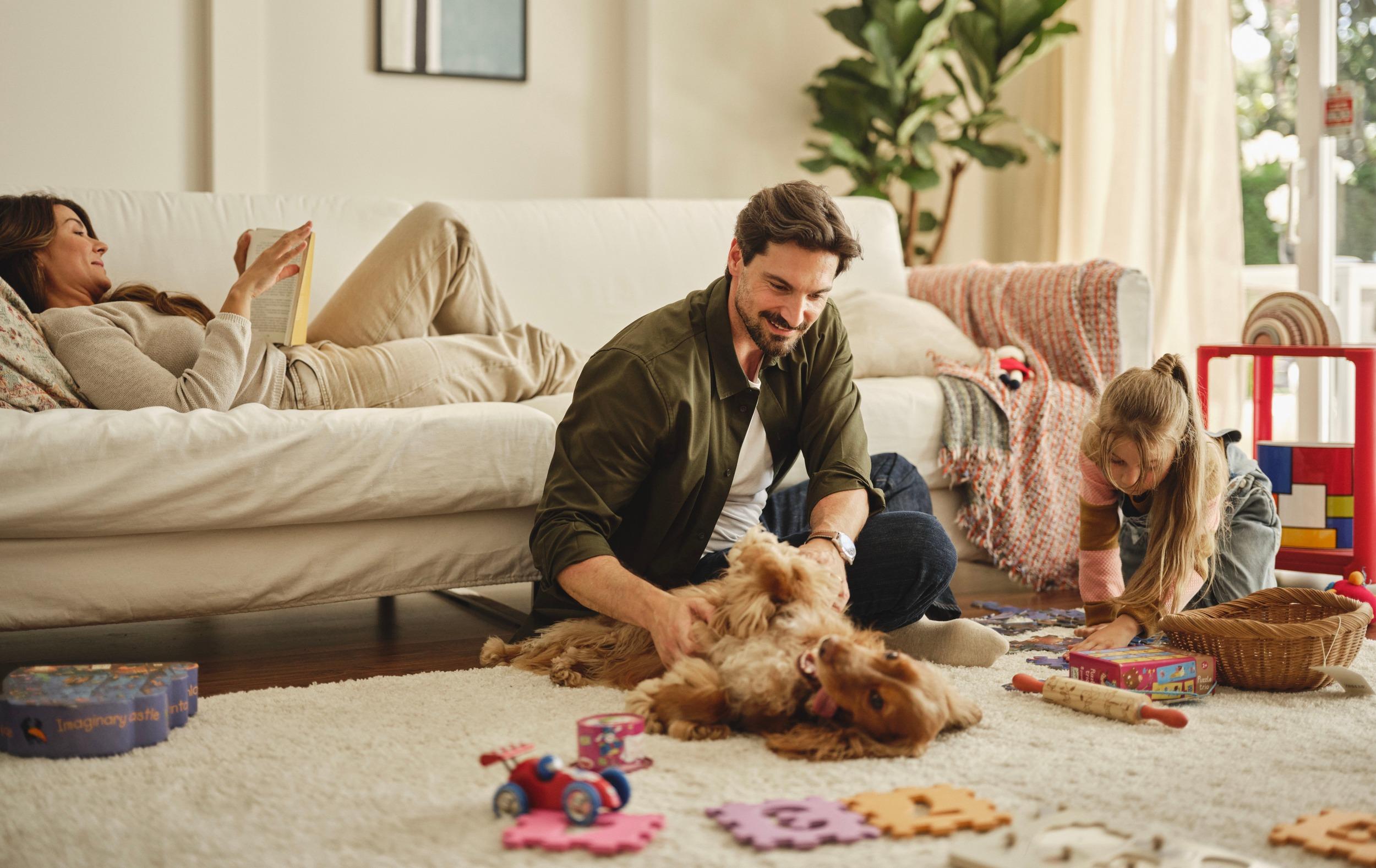 Man plays with dog while child does puzzles and woman reads on sofa.